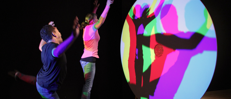 Two people dance in front of a light splitter, casting multi-coloured shadows on the wall